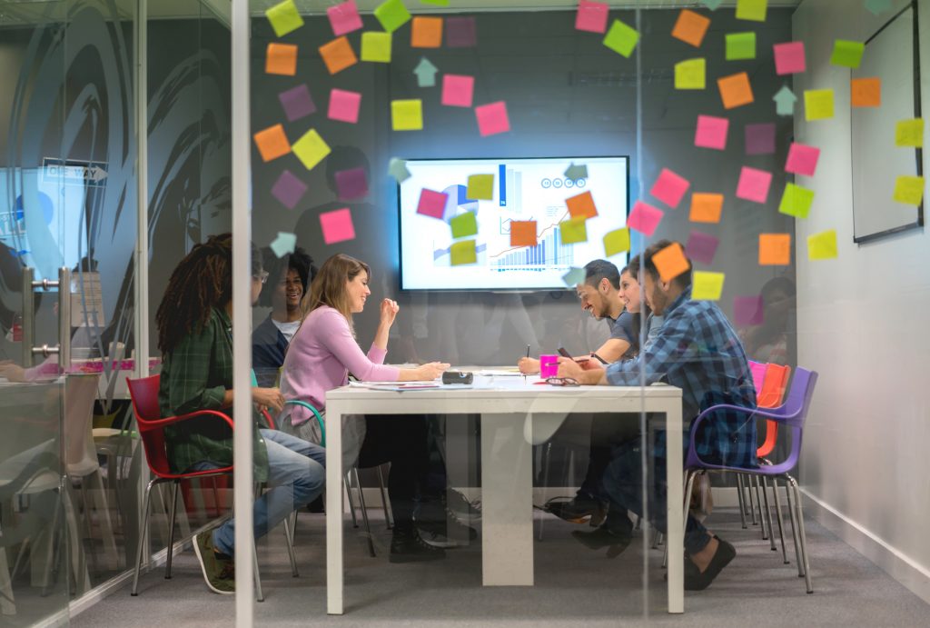 marketing professionals discuss ideas at a table, post-it notes are scattered about the office wall, a graph on a television shows an upward trend