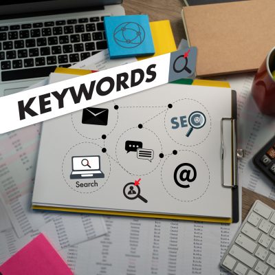 Desktop with scattered marketing notes and a clipboard, the term "keywords" floats digitally over the top inside a search bar.